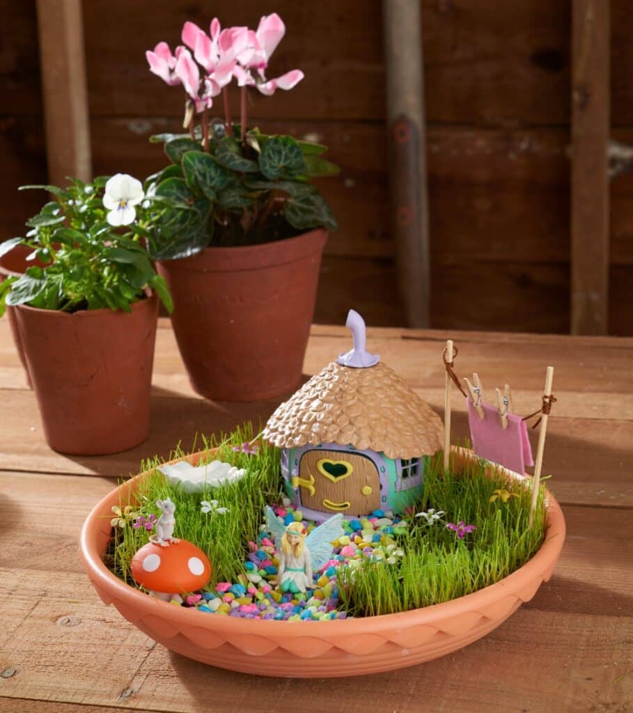 A shallow terracotta dish holds a My Fairy Garden scene—complete with a tiny house, colourful pebbles, artificial grass, a fairy figure, and clothes line—inviting you to play and discover the magic of nature amid potted plants on a wooden table.
