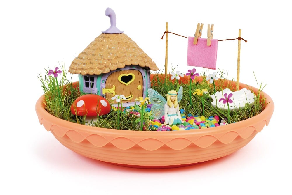 A round orange dish holds a miniature My Fairy Garden, where grass, colourful pebbles, a small fairy figure, a tiny house, and vibrant flowers invite you to play and discover the magic of nature in every charming detail.
