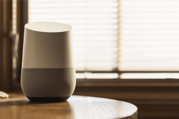 A smart speaker with a white top and grey fabric base sits on a wooden table in front of a window with closed blinds, lit by soft natural light—bringing the convenience of smart home technology to your space.