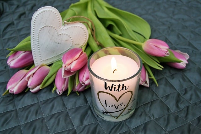 A lit candle in a glass holder with "With love" sits beside pink tulips and a white heart on a quilted grey surface—thoughtful remembrance gifts to honour loved ones.