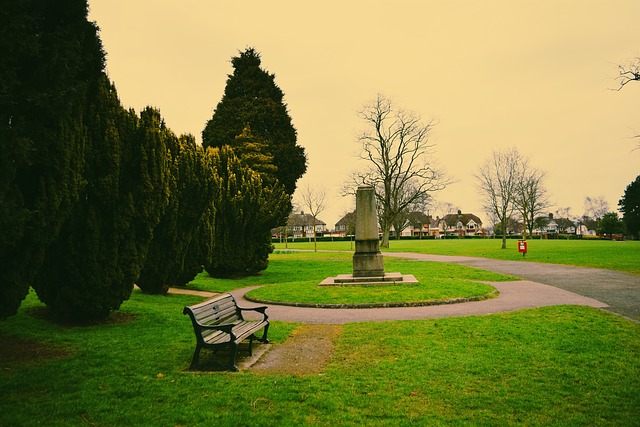 A park with a curved path, a bench, and a stone monument—perfect for reflection and remembrance gifts—sits among grass and trees. Houses are visible in the background under an overcast sky.