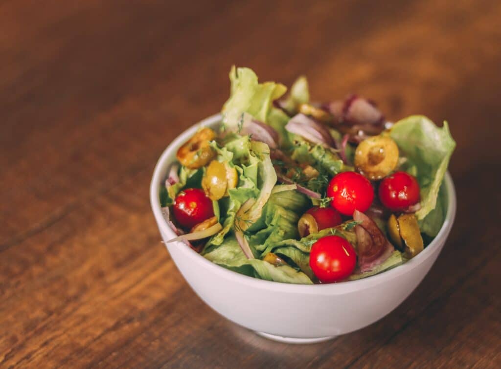 A white bowl filled with a fresh salad of lettuce, cherry tomatoes, sliced green olives, and red onion sits on a wooden table—perfect for those seeking healthy meals.