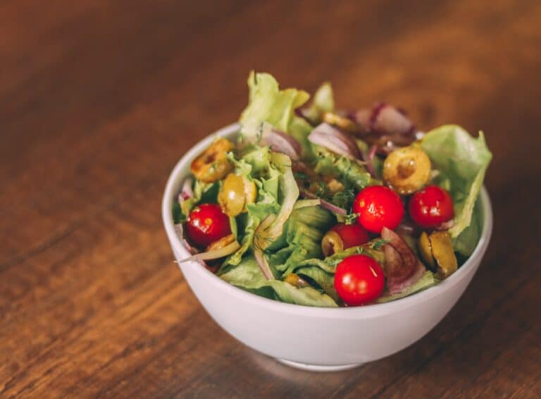 A white bowl filled with a fresh salad of lettuce, cherry tomatoes, sliced green olives, and red onion sits on a wooden table—perfect for those seeking healthy meals.