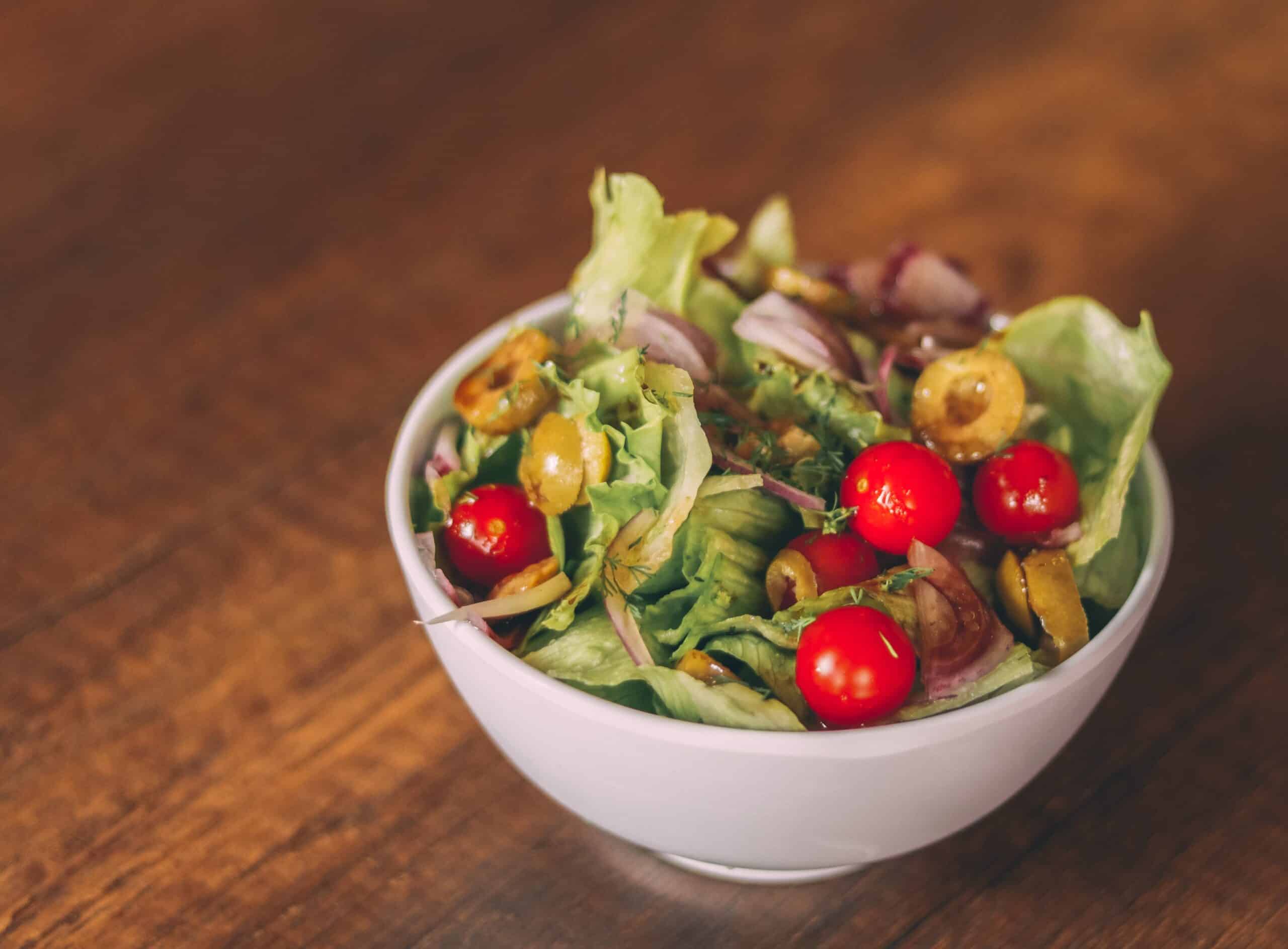 A white bowl filled with a fresh salad of lettuce, cherry tomatoes, sliced green olives, and red onion sits on a wooden table—perfect for those seeking healthy meals.
