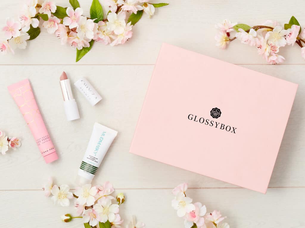 A pink GLOSSYBOX sits on a white wooden surface, surrounded by spring flowers—a perfect addition to your 2018 Mother’s Day Gift List. Beside it are a pink tube, a nude lipstick, a small white box, and a white tube of skincare product.