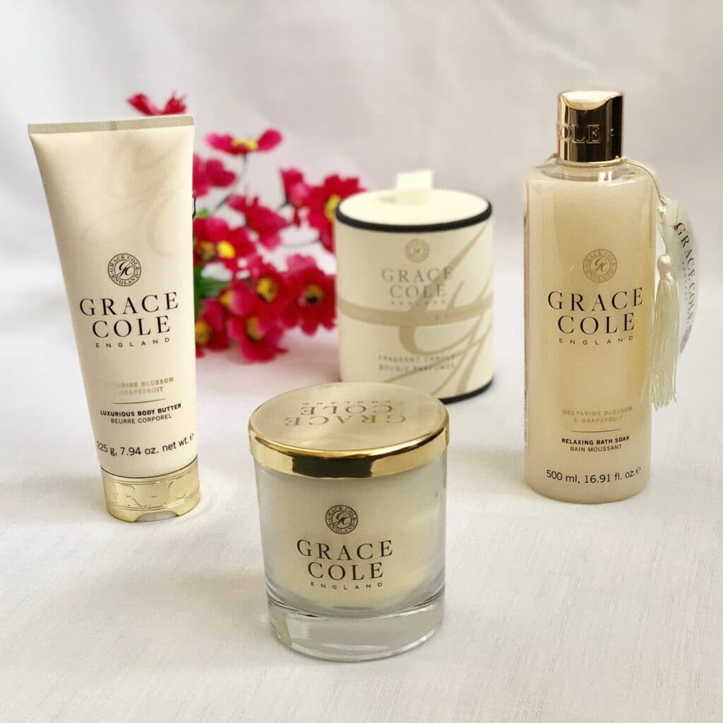 A collection of Grace Cole England products, including body butter, a candle, and a bath soak, displayed on a white surface with pink flowers—perfect for your Mother’s Day 2018 gift list.