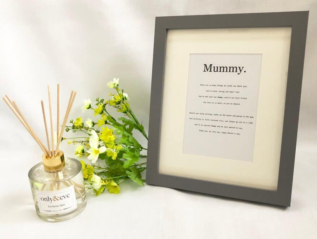 A framed print with the word "Mummy" and a sweet poem sits on a white surface beside yellow flowers and a glass reed diffuser—an ideal addition to any Mother's Day Gift List.