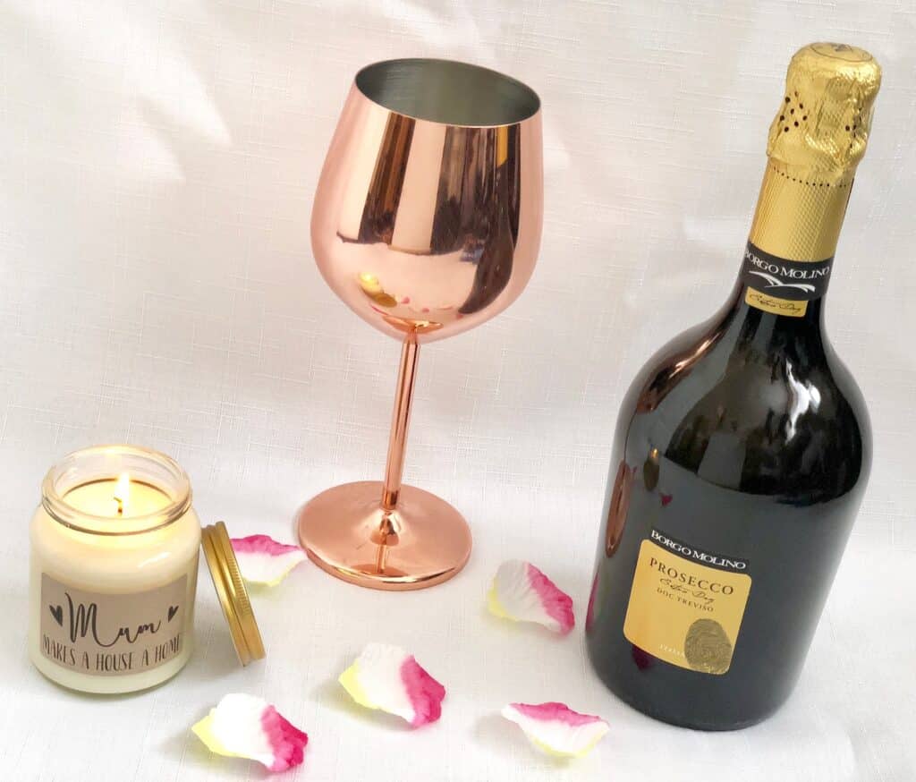 A perfect Mother’s Day gift: a lit candle in a glass jar labelled "Mum makes a house a home," paired with a rose gold wine glass, 2018 Prosecco, and scattered pink and white rose petals on a white background.