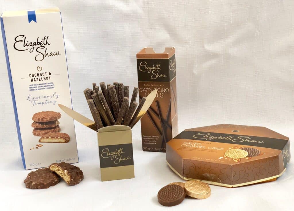 A selection of Elizabeth Shaw chocolate products, perfect for a Mother’s Day Gift List 2018: Coconut & Hazelnut biscuits, dark chocolate cappuccino flutes, and a box of salted caramel crisp chocolates on a white background.