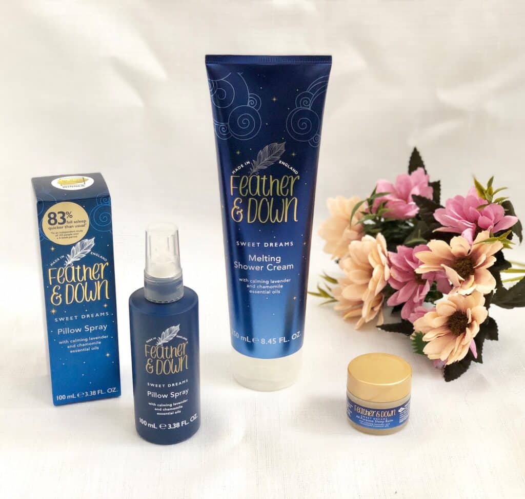 Four Feather & Down Sweet Dreams products—a pillow spray, shower cream, and balm—are displayed with their packaging beside a bouquet of pink and cream artificial flowers. Perfect for any Mother’s Day 2018 gift list.
