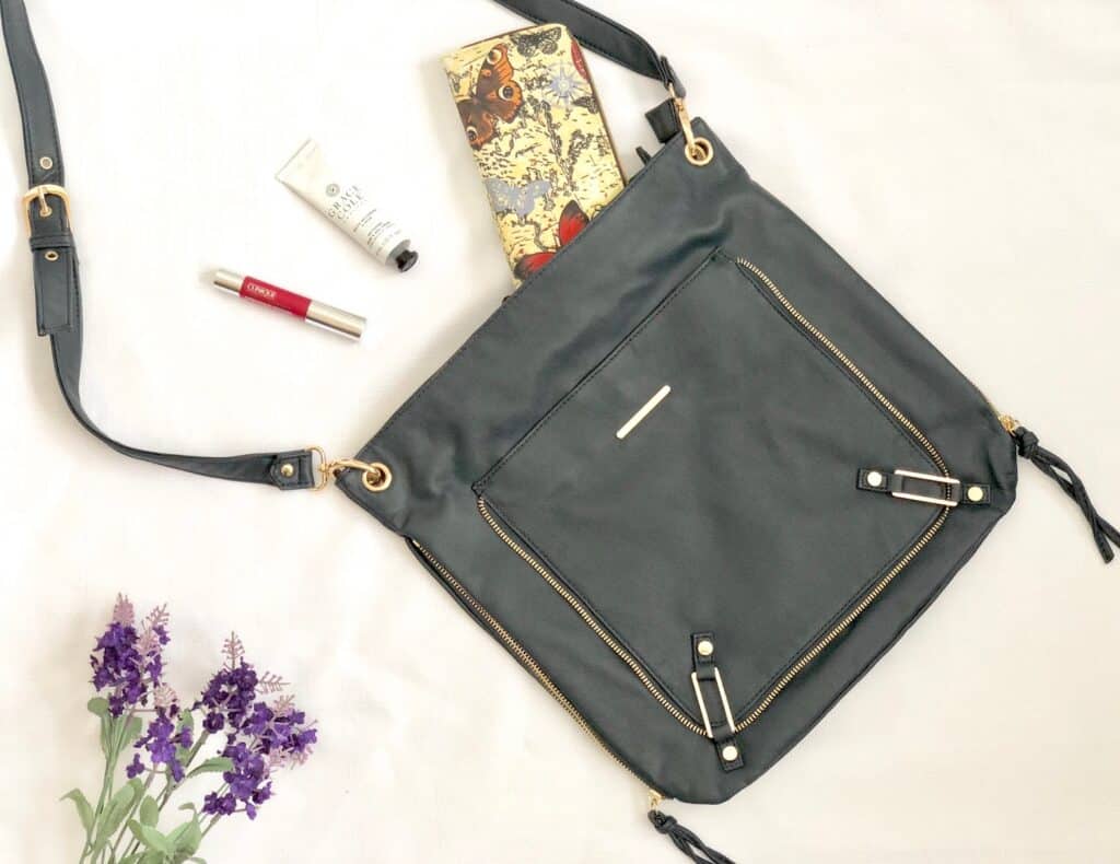 A black crossbody bag with gold zips is laid flat, its contents—floral purse, red lipstick, hand cream—displayed beside purple flowers. Perfect for your Mother’s Day gift list or as a stylish 2018 accessory.
