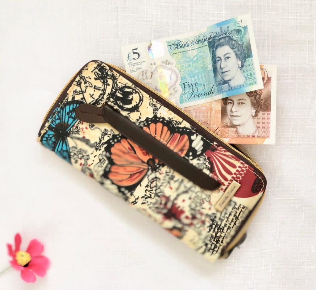 A patterned wallet with colourful butterflies rests on a white surface—an ideal choice for Mother’s Day gifts. Behind it are two British banknotes—a blue £5 note and a brown £10 note. A pink flower lies nearby.