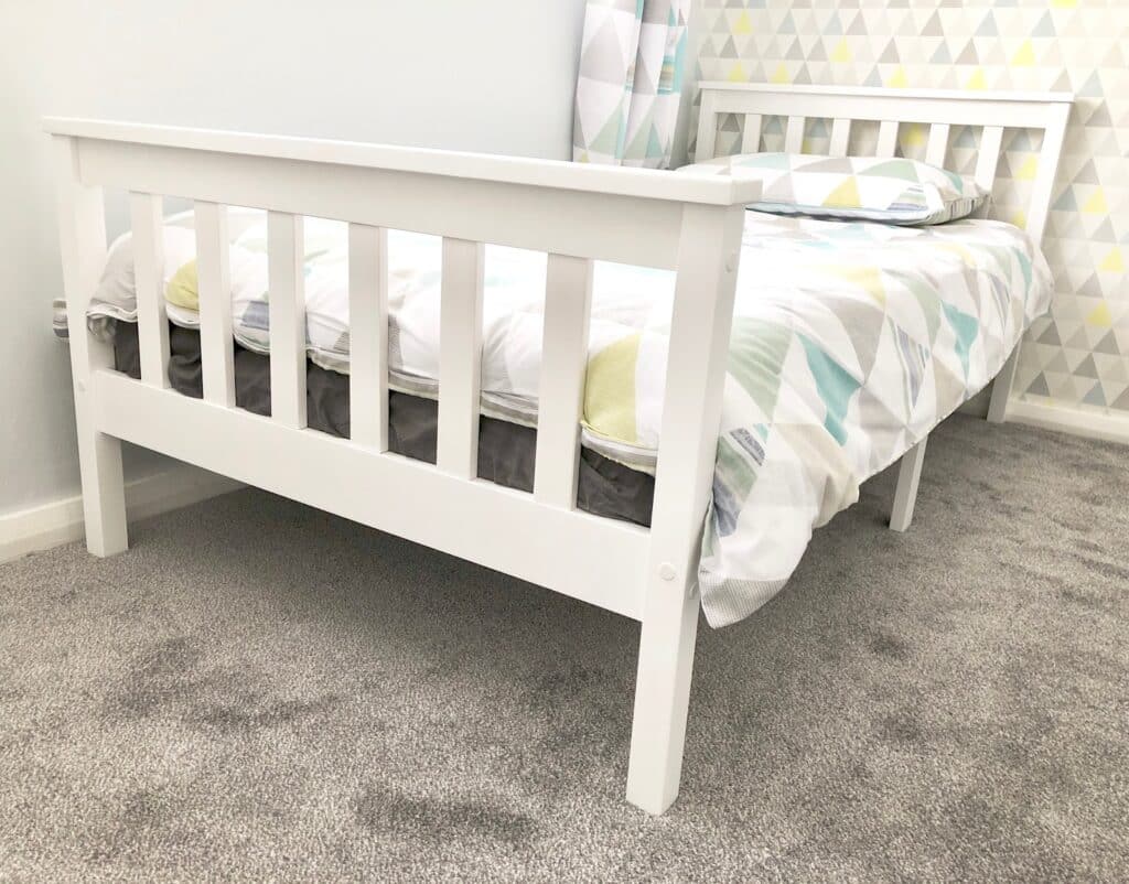 A white wooden bed frame with a geometric-patterned duvet and pillow sits on a light grey carpet in a modern boys' bedroom, featuring stylish wallpaper and a bedroom design that’s perfect for growing with ages.