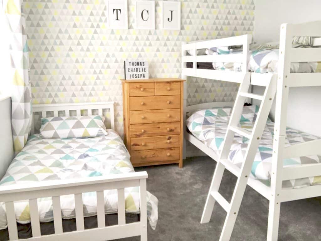 A bright boys' bedroom featuring a white single bed, wooden chest of drawers, and bunk beds. The grey carpet and geometric triangle-patterned wallpaper in pastel colours add to the playful kids' room decor.