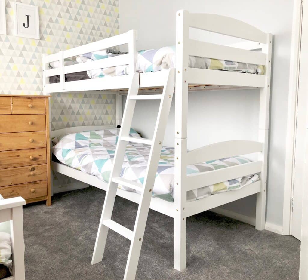 A white wooden bunk bed with patterned bedding stands in a carpeted boys’ bedroom. A wooden chest of drawers is to the left, and the wall behind features geometric triangle wallpaper and a framed letter "J," offering fresh kids’ room decor inspiration.