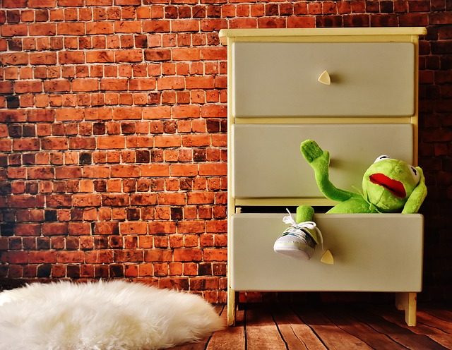 A green Kermit the Frog soft toy is partially inside an open chest of drawers, one leg and arm hanging out. The chest stands on a wooden floor against a brick wall—a playful reminder of repurposing old furniture in fun ways.