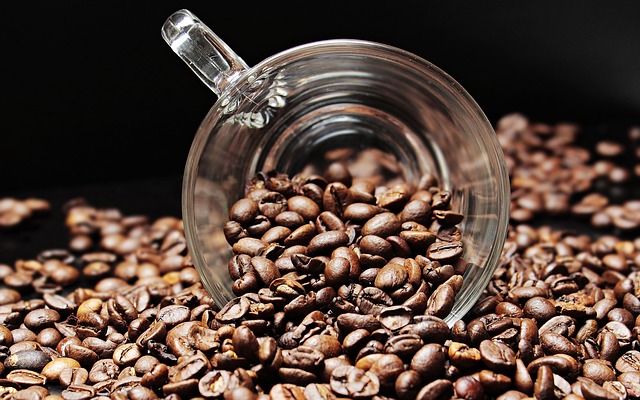 A clear glass coffee cup tipped over on its side, spilling out dark roasted coffee beans—like saving money on everyday costs, each bean adds up—onto a surface covered with more beans, against a dark background.