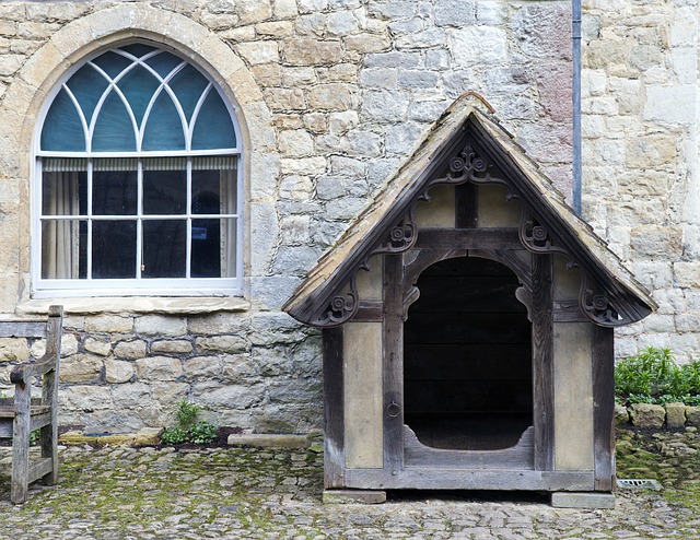 A large, ornate wooden kennel stands on a cobbled ground beside an old stone building with a tall, arched window—an unexpected glimpse of style that reflects how millennials renting seek unique charm in every corner.