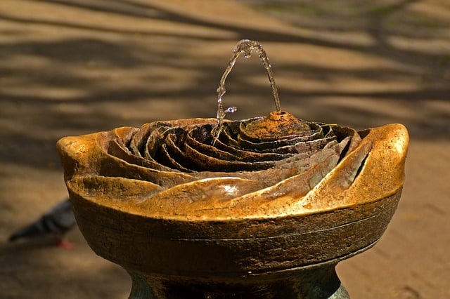 A close-up of a bronze drinking fountain shaped like a flower, with water arcing up and splashing down onto layered petals—an inspiring garden design feature, set outdoors on a sunny day.