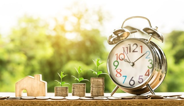 A small wooden house, three stacks of coins with seedlings growing on top, and a silver alarm clock on a table symbolise how saving money and reducing everyday costs can lead to financial growth over time.