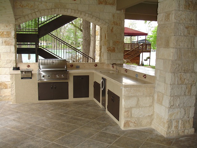 Outdoor kitchen with stone walls, built-in stainless steel grill, sink, and storage cupboards; nestled under an archway near a staircase. Overlooking a lake and trees, this space is a true garden haven for outdoor dining.