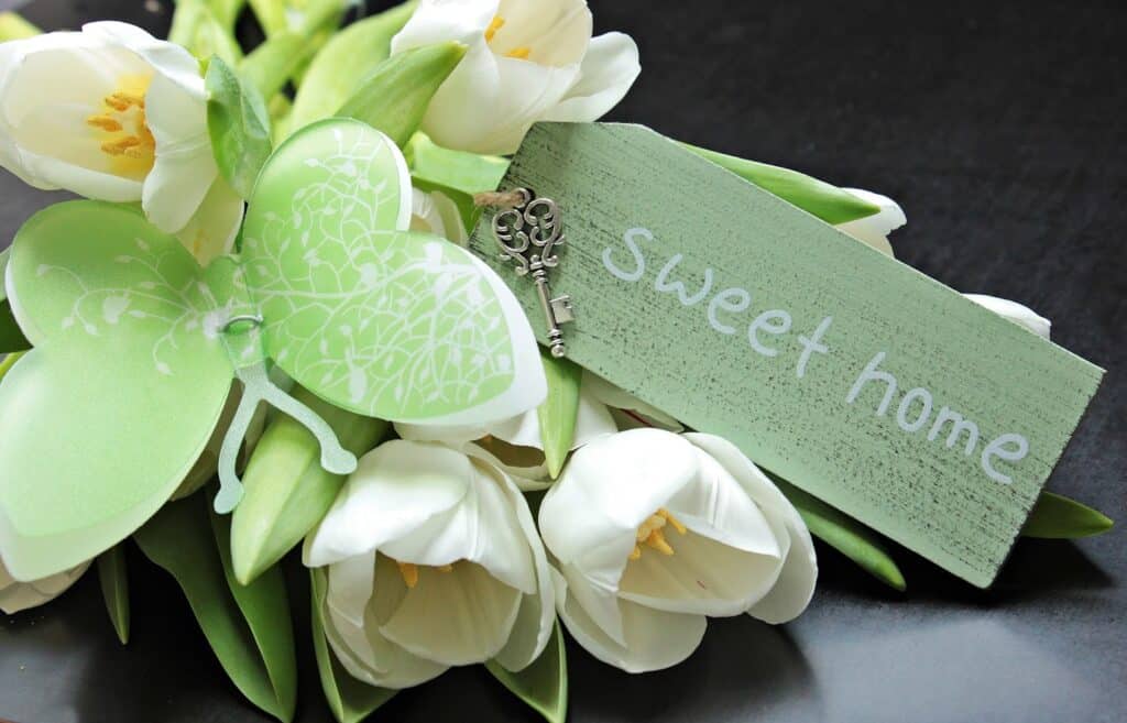 A bouquet of white tulips with a green butterfly decoration, a small vintage key, and a green wooden tag that reads "sweet home" offers charming home improvement inspiration on a dark background.