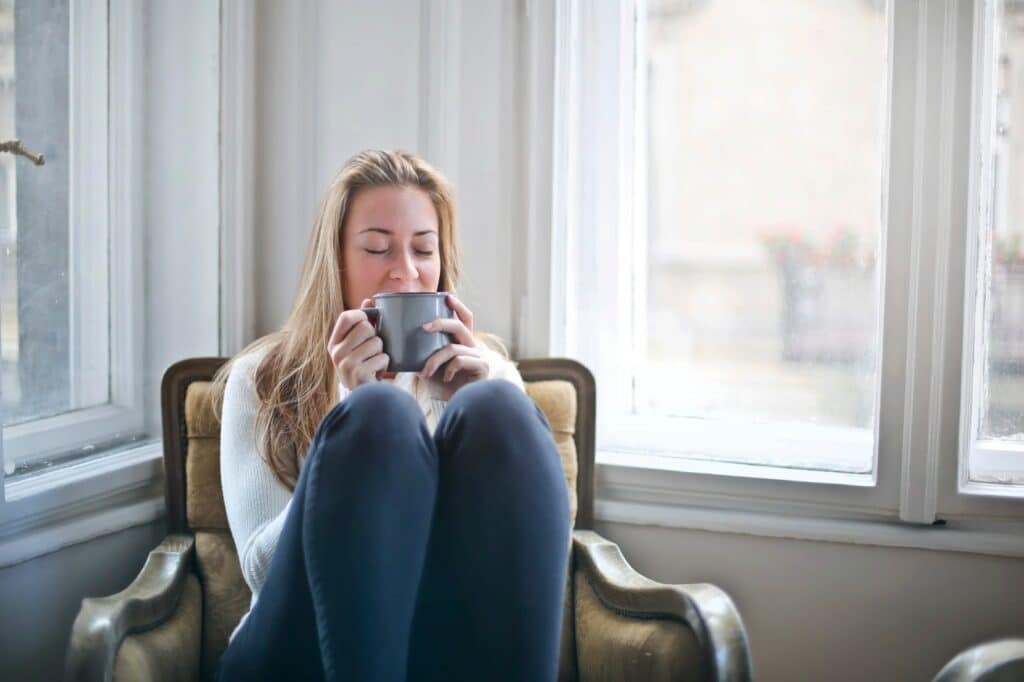 A woman sits in an armchair by large windows, holding a mug close to her face with her eyes closed, appearing relaxed and content—proof that when you love your home, every moment becomes more peaceful. She is dressed casually in a light jumper and dark trousers.