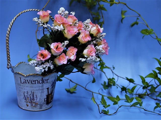 A decorative metal bucket labelled "Lavender" holds a bouquet of pink roses and white baby's-breath flowers, showcasing how artificial plants can look cool and vibrant against a blue background with green vines trailing around.