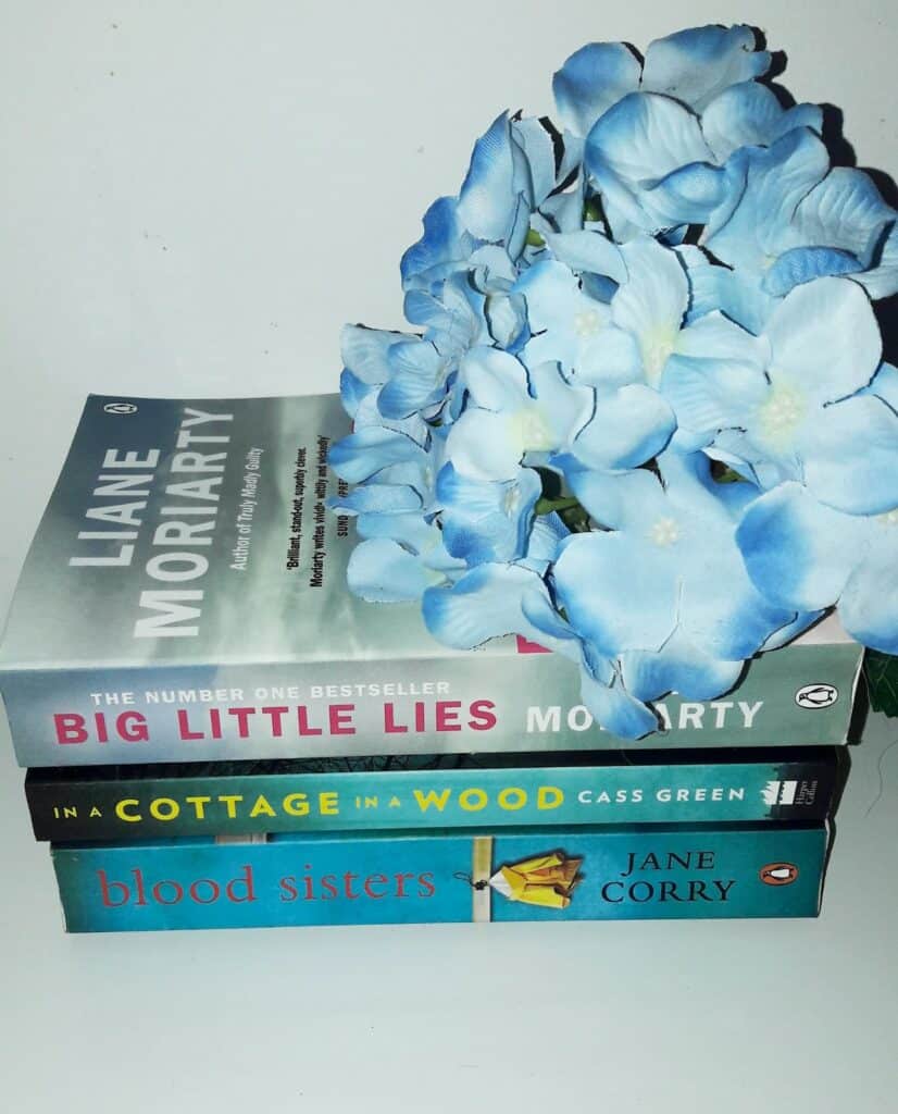 A stack of three books—“Big Little Lies,” “In a Cottage in a Wood,” and “Blood Sisters”—with blue artificial flowers on top, perfect for some Me Time, set against a plain white background. Guest Post by Joanna Victoria.