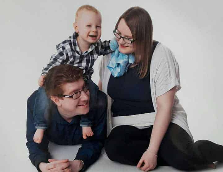 A smiling family poses against a white background. A young boy in a checked shirt sits on a man's back, both smiling, while Joanna Victoria, wearing glasses, sits beside them, looking at the boy affectionately—capturing the essence of joyful self-care moments.