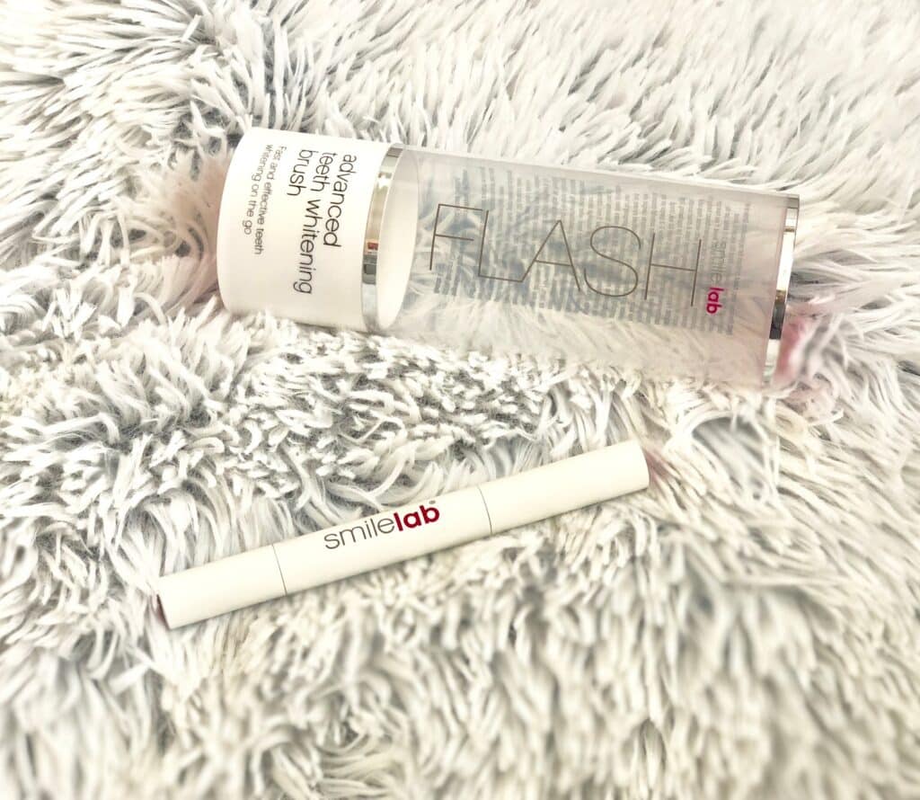 A tube of SmileLab FLASH lab advanced teeth whitening brush and pen, perfect for pre-holiday beauty essentials, lies on a fluffy white textured surface, with the pen placed below the cylindrical product container.