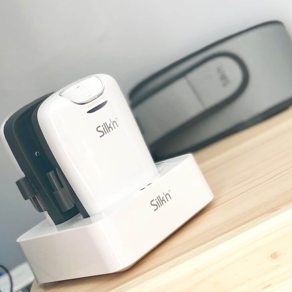 A close-up of a white Silk'n Lipo hair removal device sitting in its charging base on a light wooden surface, with another Silkn device blurred in the background—perfect for combating body issues.