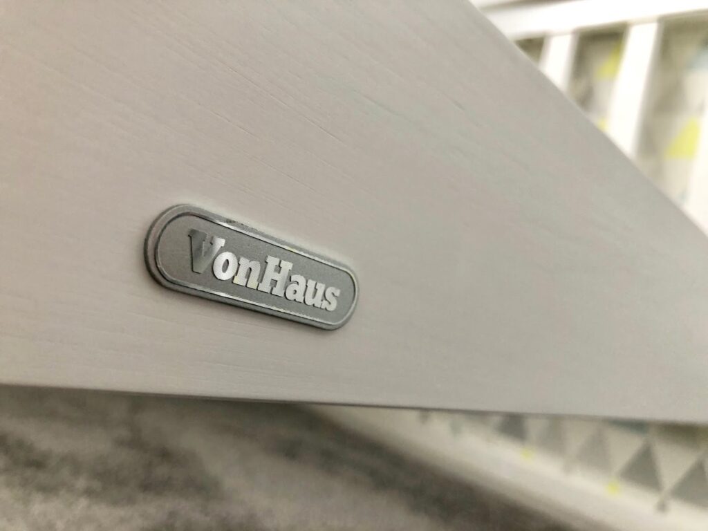 Close-up of a white wooden surface with a silver "VonHaus" logo plaque attached. In the blurred background, part of a geometric-patterned object is visible, hinting at the sturdy design perfect for any Single Pine Bed.