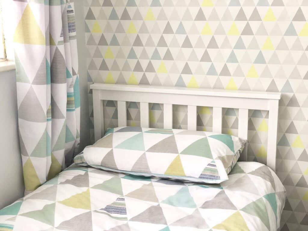 A neatly made VonHaus Single Pine Bed with a white frame, matching geometric-patterned bedding, and pillow. The wall and curtain feature pastel triangle designs in white, grey, yellow, and mint green. Light from a window is visible.