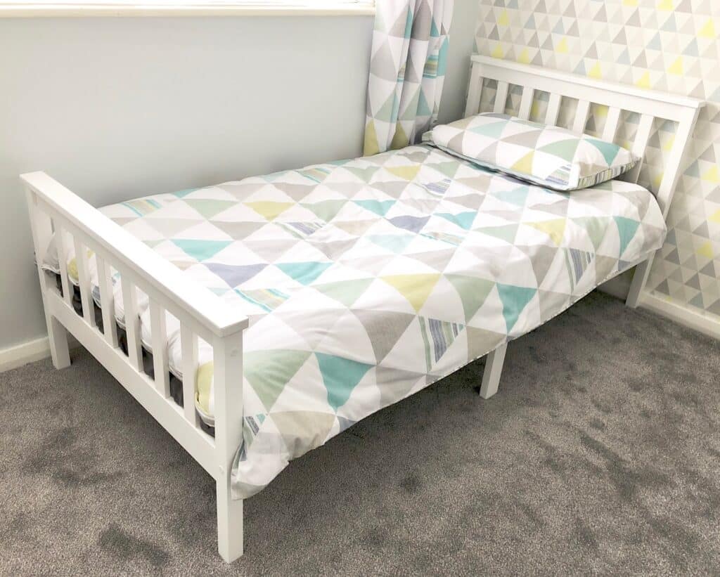 A white VonHaus Single Pine Bed with a geometric-patterned duvet and pillow in pastel colours sits on a grey carpet, next to a wall with triangle-patterned wallpaper and near a window with matching curtains.