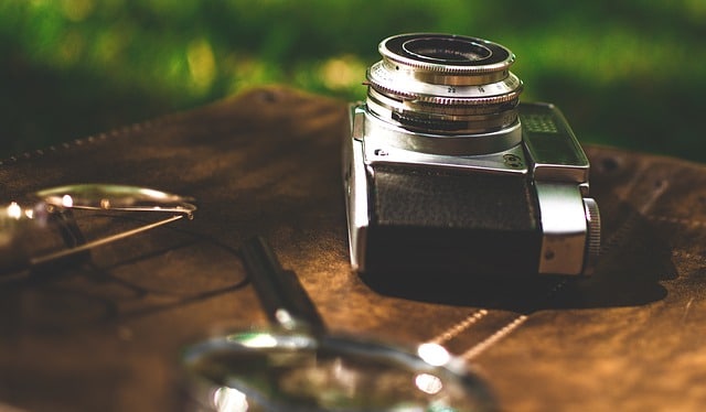 A vintage film camera rests on a brown leather surface outdoors, hinting at endless photography opportunities, with part of a pair of spectacles and green grass blurred in the background.