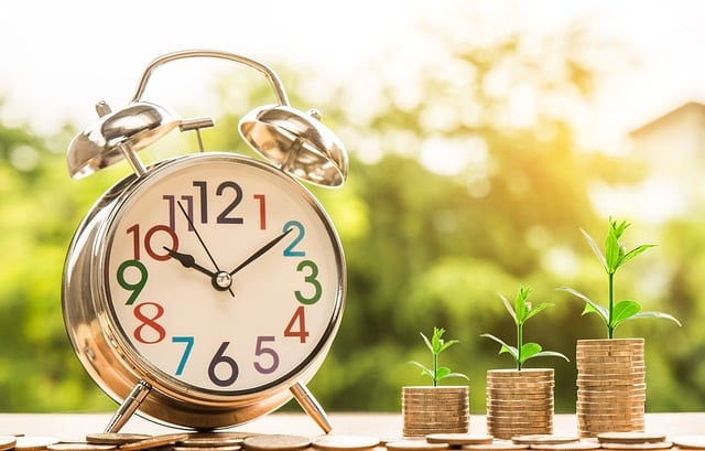 A silver alarm clock sits next to three stacks of coins, each topped with a small green plant, symbolising how the best investment grows over time. The blurred greenery and sunlight in the background convey hope and prosperity for your family.