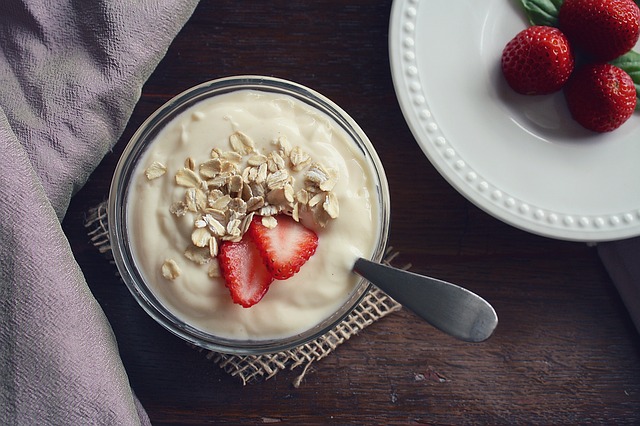 A bowl of yoghurt topped with sliced strawberries and oats sits on a small mat with a spoon inside. Nearby, a white plate holds three whole strawberries. Wondering what you should eat for a healthy smile? This wholesome snack is a great choice.