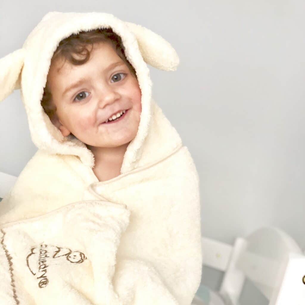 A young child with curly hair, smiling and wrapped in a soft, cream-coloured hooded towel with animal ears—one of the must-have children's holiday essentials—sits indoors against a light background.