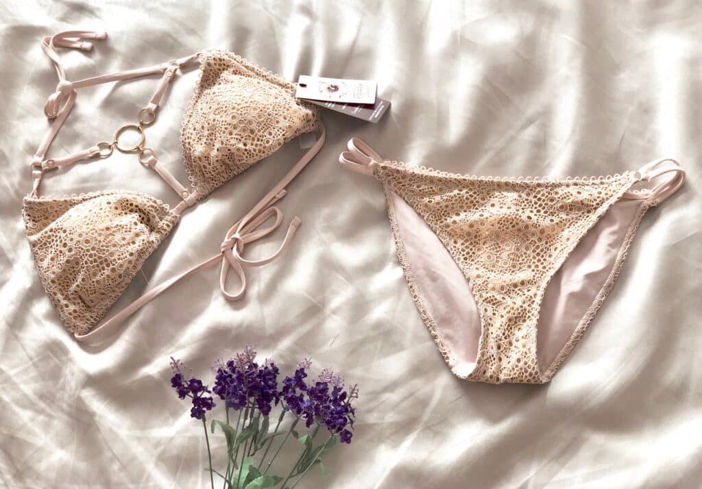 A beige lace bikini set with a gold ring detail and side ties, laid out on a white bedspread beside purple flowers—perfect for gaining body confidence this summer.