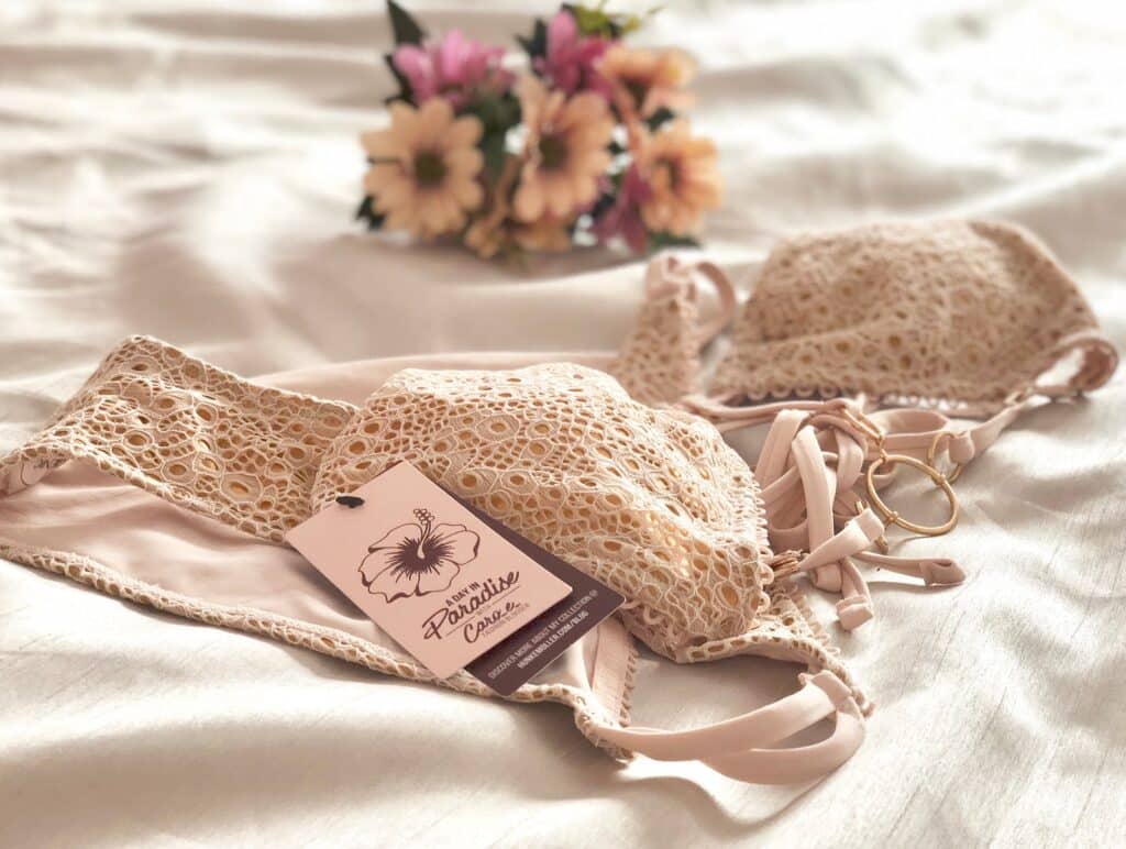 A beige lace bikini set with a "Swimwear Classic" tag is laid out on a white bedspread. In the background, a bouquet of pink and yellow flowers adds charm—perfect for inspiring gaining body confidence this season.