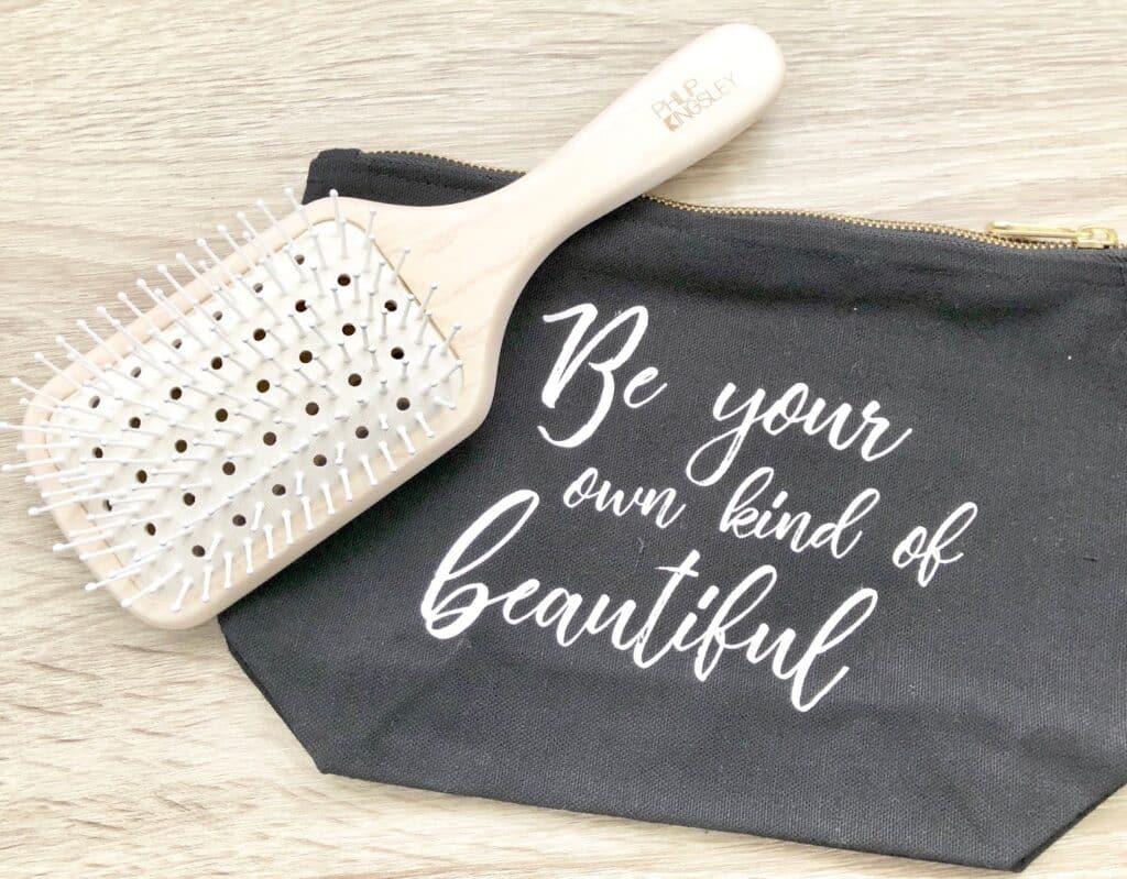 A wooden hairbrush, ideal for hair maintenance and caring for bleached hair, rests on a black zippered pouch with white script lettering that reads, "Be your own kind of beautiful." The items are placed on a light wood surface.