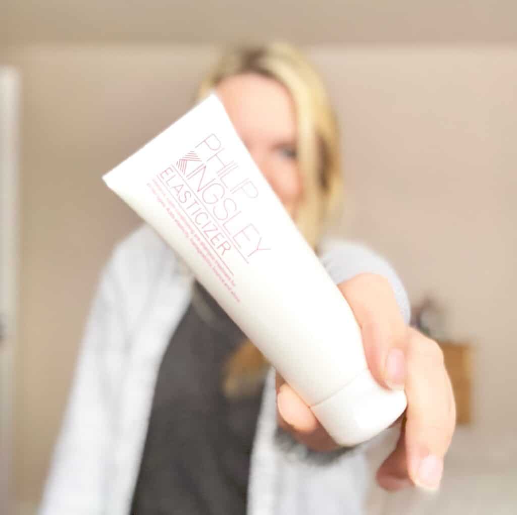 A person with blonde, bleached hair holds a tube of Philip Kingsley Elasticiser hair treatment towards the camera, highlighting this essential hair maintenance product against a softly blurred background.