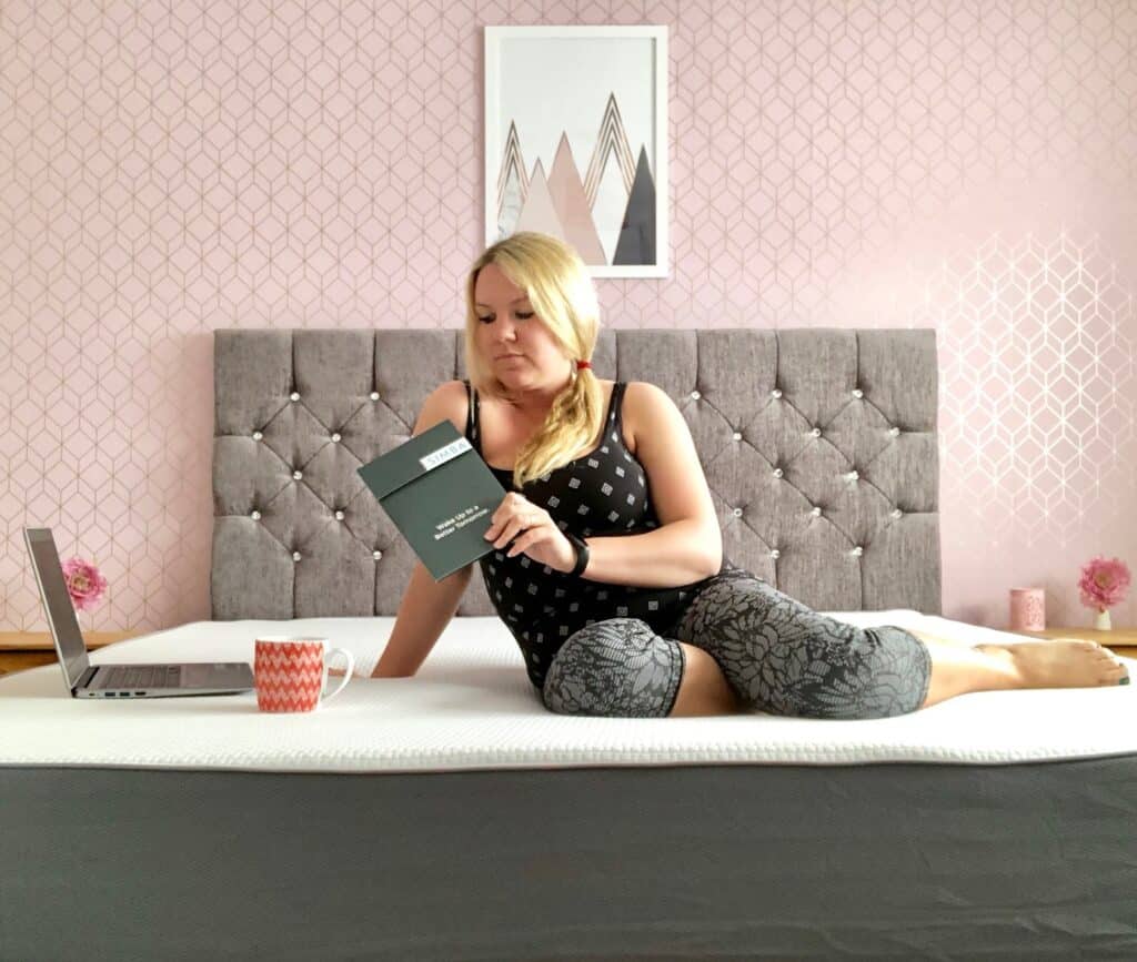 A woman with blonde hair sits on a bed with a grey tufted headboard, reading a book. A laptop and red mug rest on her Simba Hybrid Super King Mattress, in a room with pink wallpaper and geometric art above the bed.