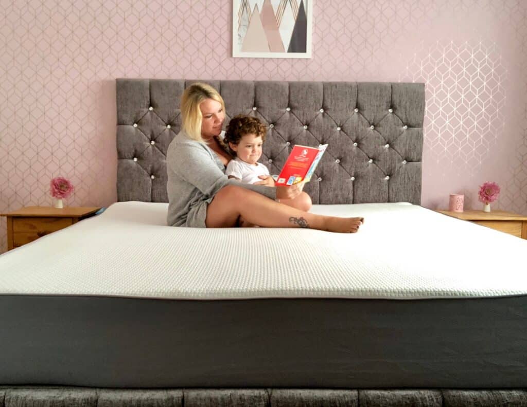 A woman and a child sit on a large Simba Hybrid super king mattress, reading a red book together. The room features pink patterned wallpaper, a grey upholstered headboard, and pink flowers on wooden bedside tables.