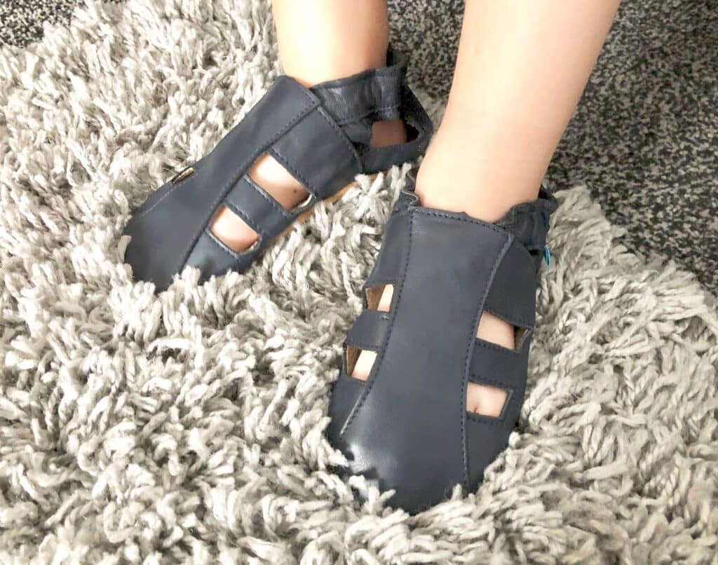 A close-up of a child's feet wearing navy blue leather sandals, perfect for children's holiday essentials, with cut-out sections, standing on a soft, light grey shaggy rug.