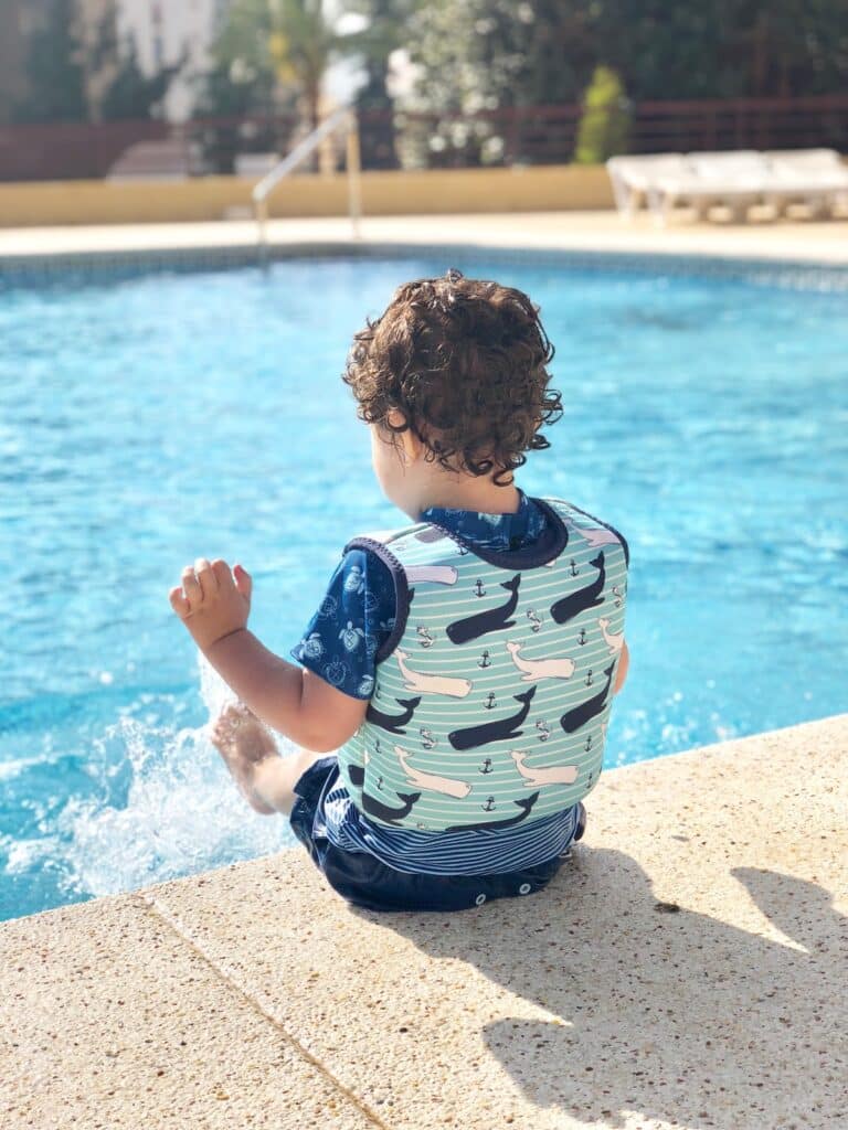 A young child with curly hair, wearing a whale-patterned swim vest and blue swim shorts—perfect children's holiday essentials—sits at the edge of a swimming pool, playfully kicking the water on a sunny day.
