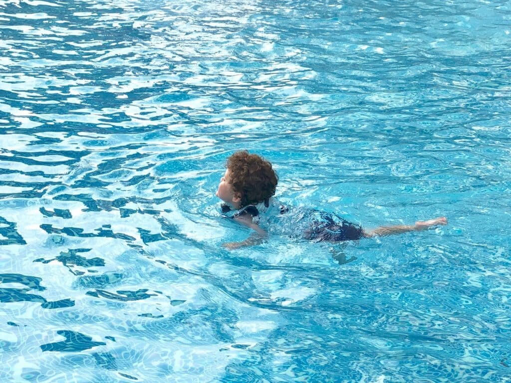 A young child with curly hair swims alone in a clear blue pool, wearing blue swimming trunks—one of the children's holiday essentials—using a swimming style that keeps their head above the water.