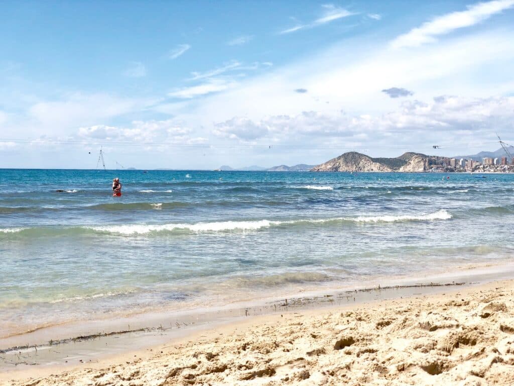 A sandy beach with gentle waves, a person standing in the shallow water, distant hills, and a city skyline under a partly cloudy blue sky—perfect for your first family holiday.