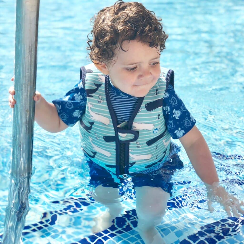 A young child with curly hair, wearing a swim vest—one of the key children's holiday essentials—and a blue swimming costume, stands on a pool step holding onto a metal rail, sunlight reflecting on the clear blue water.